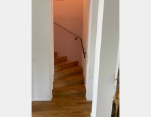 Holztreppe in einem Wohnbereich, die in einer Ecke nach oben führt, mit weißen Wänden und einem schwarzen Handlauf an der rechten Seite. Der Boden besteht aus hellem Holzparkett.