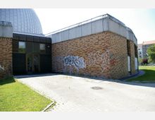 Backsteinbau mit flachen rechteckigen Fenstern und einer runden Metallkuppel im Hintergrund. Eine mit Graffiti bedeckte Ziegelwand sowie ein gepflasterter Vorplatz grenzen an eine Grünfläche.