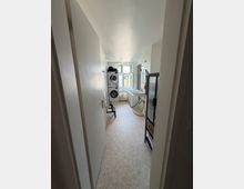 Schmaler Hauswirtschafts- und Waschraum mit weiß gefliesten Wänden, hellem Boden und einem Fenster am Ende. Links stehen übereinander Waschmaschine und Trockner, rechts sind Waschbecken, Badewanne und ein Duschbereich mit Glasabtrennung angeordnet.