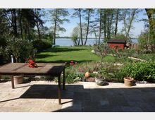 Terrasse mit Steinfliesen und Holztisch, im Hintergrund ein gepflegter Garten mit Blumenbeeten, Rasenfläche und Bäumen, dahinter ein See und ein rotes Gartenhaus am Rand.