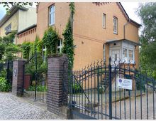 Zweistöckiges Backsteingebäude mit hohen Fenstern und Kletterpflanzen an der Fassade, umgeben von einem schwarzen, kunstvoll verzierten Metallzaun mit einem Tor aus Backsteinpfeilern. Der Eingang verfügt über einen weiß gestrichenen Erker und eine gepflasterte Einfahrt.