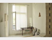 Ein minimalistisches Schlafzimmer mit weiß gestrichenen Wänden, einem großen Fenster mit halb geöffnetem Vorhang, einem kleinen Wandtisch mit einem Spiegel, einer Vase und Büchern darauf. Daneben stehen Bücherstapel, ein grüner Hocker und ein schlichtes Bett.