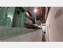 Industrielle Innenansicht mit einer großen grünen Maschinenanlage auf einer massiven weißen Betonbasis. Die Umgebung besteht aus Betonwänden und einer zum Teil sichtbaren Holzdecke mit Metallstreben.