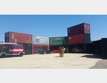 Ein offener Platz mit gestapelten Schiffscontainern, die als Lagerräume oder Werkstätten genutzt werden. Im Vordergrund steht ein rotes Feuerwehrfahrzeug sowie ein schwarzer Oldtimer, daneben ein Baumstumpf und einige rote Fässer.