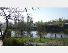 Parklandschaft mit einem Fluss im Vordergrund, umgeben von Bäumen und Sträuchern. Ein Pfad mit einer Holzbank verläuft entlang des Wassers, im Hintergrund eine kleine Brücke und grasbewachsene Hügel.