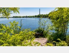 Ufer eines Sees mit dichter, grüner Vegetation im Vordergrund und einem schmalen Sandweg, der zum Wasser führt; im Hintergrund sind ein Industriekamin und weitere Bäume sichtbar.