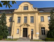 Gelbes, zweistöckiges Gutshaus mit barocken Elementen, wie einem geschwungenen Giebel und verzierten Fenstern. Vor dem Gebäude stehen zwei Nadelbäume, ein weißer Gartenstuhl, sowie ein Schild mit der Aufschrift "Gutshaus".