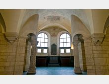 Innenraum einer romanischen Krypta mit dicken steinernen Pfeilern, Rundbögen und Kreuzgewölbe. Im Hintergrund befinden sich große Fenster mit darüberliegenden Wandmalereien und eine zentrale Gedenktafel.