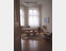 Minimalist living room with tall bay windows covered by sheer white curtains, a low sofa, and a round wooden coffee table on layered shag rugs over wide plank wood flooring. A wall-mounted TV with a small console sits on the right wall beneath a large hanging fabric pendant light.