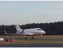 Ein kleines Passagierflugzeug rollt auf einer asphaltierten Landebahn eines Flughafens, umgeben von einer Wiese und einem Wald im Hintergrund. Im Vordergrund befindet sich ein Schild mit der Aufschrift "C 25-07".