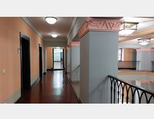 Ein heller Innenflur mit pastellfarbenen Wänden in Orange und Grau, durchzogen von rosa verzierten Kapitellen an den Säulen. Der Boden besteht aus glänzendem braunem Holz, und schwarze Metallgeländer begrenzen die offenen Bereiche neben dem Flur.