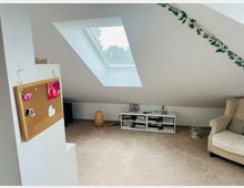 Wohnzimmer oder Arbeitszimmer im Dachgeschoss mit schräger Decke und Dachfenster, das viel Tageslicht hereinlässt. Der Raum enthält eine Korkwand mit kleinen Notizen und Fotos, ein offenes Regal mit Büchern und Dekorationen, sowie einen beigefarbenen Sessel und eine Lichterkette aus künstlichen Blättern an der Wand.