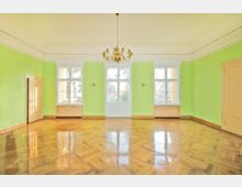 Ein heller Raum mit grünen Wänden, glänzendem Fischgrätparkettboden und großen Fenstern. In der Mitte hängt ein goldener Kronleuchter, seitlich befinden sich Holztüren und schlichte Wandlampen.