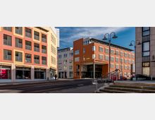 Straßenszene mit modernen Büro- und Geschäftsgebäuden, darunter ein orangefarbenes Gebäude mit der Aufschrift „Fürstengalerie“ und „Amtsgericht“. Im Vordergrund befinden sich Bürgersteige, Treppen und Straßenlaternen.