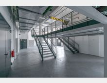 Industriehalle mit moderner Zwischengeschossebene, ausgestattet mit Metalltreppen und Geländern. Raum mit Betonboden, weiß gestrichenen Wänden und sichtbarer Deckenstruktur mit Rohren und Belüftungselementen.