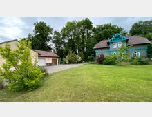 Ein weitläufiger Garten mit einem Fachwerkhaus in Blau und Braun auf der rechten Seite, umgeben von Bäumen. Links steht ein separates Gebäude mit Garagentor und rotem Ziegeldach, davor eine gepflasterte Einfahrt.