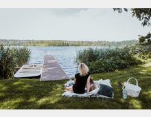 Eine Frau sitzt auf einer Decke neben einem Hund auf einer Wiese am Seeufer. Im Hintergrund befindet sich ein hölzerner Steg, der ins Wasser führt, daneben liegt ein Ruderboot, umgeben von Schilf und Bäumen.