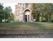 Backstein-Gebäude mit einem großen, bogenförmigen Eingangsportal, flankiert von Säulen; davor eine Rasenfläche mit einigen Bäumen im Hintergrund. Ein Schaukasten ist rechts neben dem Eingang sichtbar, und eine niedrige Metallbegrenzung säumt den Vordergrund.