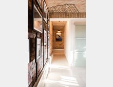 Flur mit freiliegenden Ziegelwänden und Gewölbedecke, dekoriert mit gerahmten Bildern auf der linken Seite und einem Poster am Ende des Gangs; der Boden ist mit hellen Fliesen ausgelegt, und auf der rechten Seite befindet sich eine Milchglaswand.