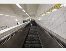 Eine Rolltreppe in einem U-Bahn-Bereich, umgeben von weißen, gefliesten Wänden mit gelbem Linienmuster. Die Treppenstufen sind mit Graffiti versehen, und mehrere Personen stehen oben auf der Rolltreppe.