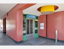 Eingang zu einer U-Bahn-Station mit einer gläsernen Automatiktür, grünem Türrahmen und Symbolen für die U-Bahnlinie U3. Die Wände sind mit roten Mosaikfliesen verkleidet, und oben hängt eine große, runde gelbe Deckenleuchte.