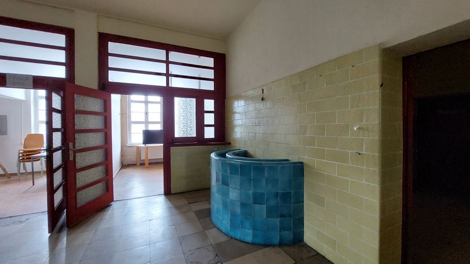 Ein Innenraum mit gelb gefliesten Wänden und einem hellblauen, halbrunden Fliesentresen. Im Hintergrund sind rote Türrahmen mit Glaselementen sowie ein Fenster mit Tageslicht zu sehen.