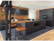 Moderne Küche mit schwarzen Schränken und einer Insel aus dunklem Material, kombiniert mit einer Holzarbeitsfläche. Die Wand ist teils aus sichtbarem Backstein und teils mit Holzvertäfelung gestaltet, ergänzt durch dunkle Fliesen und Holzboden.