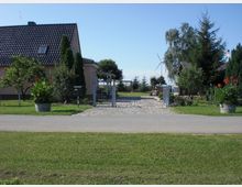 Einfamilienhaus mit dunklem Ziegeldach und umliegendem Garten, eingezäunt von einem Metalltor und dekoriert mit Blumenkübeln. Im Hintergrund sind Bäume und Windkraftanlagen auf freiem Feld zu sehen.