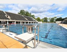 Freibad mit einem großen Schwimmbecken, Startblöcken entlang des Beckenrandes und einer Tribüne mit Sitzplätzen im Hintergrund. Im Außenbereich sind Bäume und eine offene, sonnige Atmosphäre sichtbar.