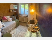 Gemütlicher Wohnraum mit warmem Licht, bestehend aus einem Tagesbett mit dekorativen Kissen, einem kleinen runden Tisch mit schwarz-weißem Muster, einem gemusterten Sessel und einer auffälligen hängenden Lampe aus Holzstreben. Die Wände sind in Kork- und Blautönen gestaltet, ein großes Fenster mit weißem Rollo und Blick ins Grüne sorgt für natürliches Licht.