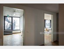 Blick durch einen Flur in zwei Räume eines modernen Büros mit Holzboden; links ein leerer Raum mit Zugang zu einer Wendeltreppe durch eine Glastür, rechts ein Konferenzraum mit Tisch und Stühlen vor großen Fenstern mit Metallstreben.