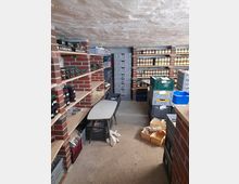 Kellerraum mit Regalen aus roten Ziegelsteinen und Holz, die voll mit Einmachgläsern und Konserven sind. Auf dem Betonboden stehen stapelbare Kunststoffkisten, ein kleiner Tisch und leere Holzkisten.