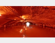 Ein unterirdischer Tunnel mit gewölbter Backsteindecke, beleuchtet durch orangefarbene Lampen. Auf dem Wasser des Kanals ist im Hintergrund ein heller Lichtschein zu sehen, der vermutlich den Ausgang markiert.
