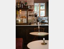 Ein kleines Café mit einer Theke aus dunklem Holz, auf der eine Kaffeemaschine, Glasglocken mit Gebäck, und Flaschen mit Getränken stehen. Im Vordergrund befinden sich runde Tische mit Kerzenhaltern, im Hintergrund sind Regale und ein Fenster mit Blick nach draußen zu sehen.