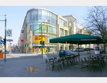 Straßenszene mit einem modernen Gebäude aus Glas und Stein, das Geschäfte beherbergt, darunter eine Filiale von "Deichmann". Im Vordergrund befinden sich leere Tische und Stühle unter einem grünen „Starbucks Coffee“-Sonnenschirm, und eine Straßenkreuzung mit Ampeln ist sichtbar.