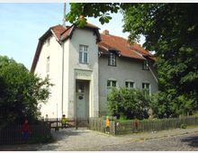 Großes, zweistöckiges Gebäude mit heller Fassade und roten Dachziegeln, gekennzeichnet als "Schulhaus". Vor dem Gebäude befindet sich ein kleiner Garten mit Sträuchern, umgeben von einem Holzzaun, und mehrere bemalte Figuren sind entlang des Zauns sichtbar.