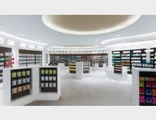 Modernes Apothekengeschäft mit heller Beleuchtung, geschwungenen Deckendesigns und weißen Regalen, die verschiedene Produkte wie Medikamente, Vitamine und Pflegeartikel präsentieren. Im Hintergrund befinden sich größere Produktregale an den Wänden, während kleinere freistehende Theken im Vordergrund stehen.