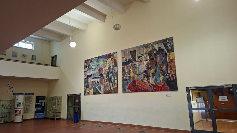 Große Innenhalle eines öffentlichen Gebäudes mit hoher Decke, sichtbaren Deckenbalken und hängenden Kugellampen. An einer hellen Wand hängen zwei großformatige, farbige Wandbilder; daneben stehen Vitrinen und Beschilderungen, rechts führt eine Glastür in einen Flur.
