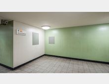 Ein heller U-Bahn-Tunnel mit gefliesten Wänden in Grün- und Weißtönen, einem Wegweiser zur Linie U9 „über den Bahnsteig“ und einer Deckenleuchte. Der Boden ist mit grauen Fliesen ausgelegt, und in einer Ecke ist eine Überwachungskamera angebracht.