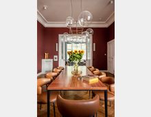 Esszimmer mit einer langen Holztischplatte, umgeben von gepolsterten braunen Stühlen. Der Raum hat weinrote Wände, eine moderne Hängelampe mit Glasglobus-Design und eine Vase mit Sonnenblumen als Tischdekoration; im Hintergrund sind weiße Doppeltüren und große Fenster erkennbar.