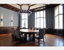 Historischer Konferenzraum mit einem großen ovalen Holztisch, umgeben von gepolsterten Stühlen. Der Raum hat hohe Fenster mit Vorhängen, holzvertäfelte Wände und eine große, moderne Deckenleuchte.