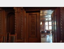 Holzvertäfelter Innenraum mit detaillierten Schnitzereien an Türrahmen und Wänden. Im Hintergrund ist ein heller Raum mit großen Fenstern, hölzernen Möbeln und hängenden Lampen zu sehen.