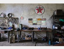 Werkstatt mit rustikaler Einrichtung: Eine lange Werkbank aus Holz mit Werkzeugen, Geräten und Autoteilen darauf; an der Wand hängen Nummernschilder und ein Texaco-Logo, daneben Regale mit weiteren Werkzeugen und Zubehör.