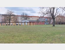 Weite Grünfläche in einem Park mit vereinzelten Bäumen, im Hintergrund mehrstöckige Wohngebäude mit teils gelben und roten Fassaden sowie ein Backsteingebäude. Eine schlichte überdachte Säulenkonstruktion erstreckt sich parallel zur Wohnbebauung.