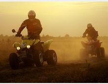 Zwei Personen fahren bei Sonnenuntergang auf staubigen Wegen Quad-Motorräder durch eine offene Landschaft. Die Szene ist in warmes, goldenes Licht getaucht, und im Hintergrund sind niedrige Vegetation und ein wolkiger Himmel zu sehen.