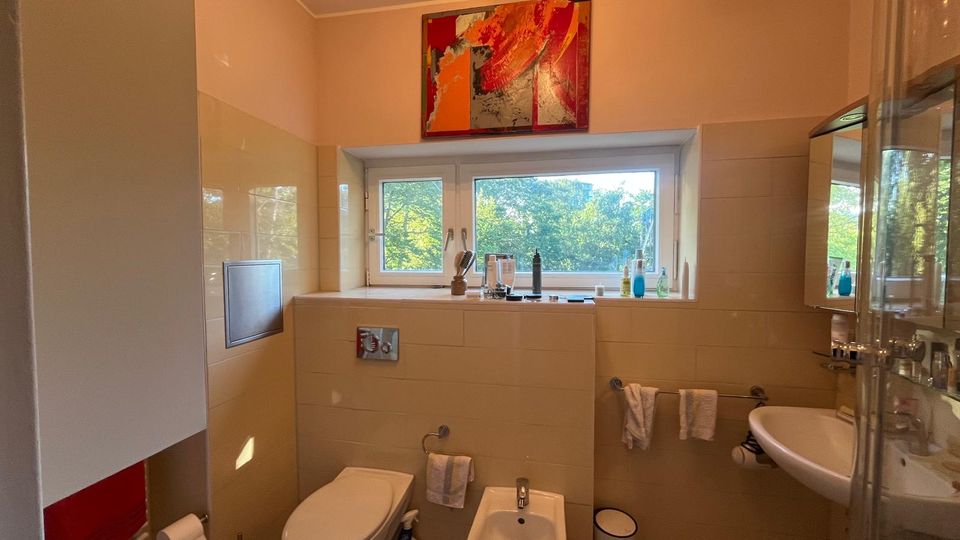 Modernes Badezimmer mit beigefarbenen Fliesen, wandhängendem WC und Bidet sowie Waschbecken neben einer Glasduschkabine. Ein breites Fenster mit Blick ins Grüne und ein abstraktes Bild darüber prägen den Raum.