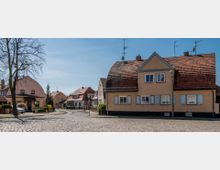 Straßenszene in einem Wohngebiet mit Einfamilienhäusern, die überwiegend beige und mit roten Ziegeldächern gestaltet sind. Der gepflasterte Straßenverlauf ist von einfachen Vorgärten, einem Baum und leicht hügeligen Häuserreihen umgeben.