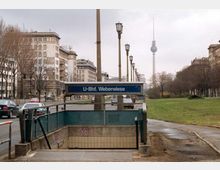 U-Bahn-Eingang „U-Bhf. Weberwiese“ an einer Stadtstraße, flankiert von mehrstöckigen Wohngebäuden und Bäumen; im Hintergrund ist der Berliner Fernsehturm sichtbar.