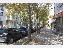 Straßenszene in einer Wohngegend mit mehrstöckigen Gebäuden auf der linken Seite und Geschäften auf der rechten Seite. Am Straßenrand stehen parkende Autos, während Bäume entlang des Gehwegs Schatten spenden; ein Schild weist auf eine Tempo-30-Zone hin.