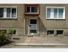 Fassade eines älteren Mehrfamilienhauses mit bräunlichem Putz, weißen Fensterrahmen und einem Eingang mit Glastür und eingelassenen Briefkästen. Vor dem Gebäude befinden sich ein kleiner gepflasterter Gehweg, ein Fahrradständer und etwas Begrünung.